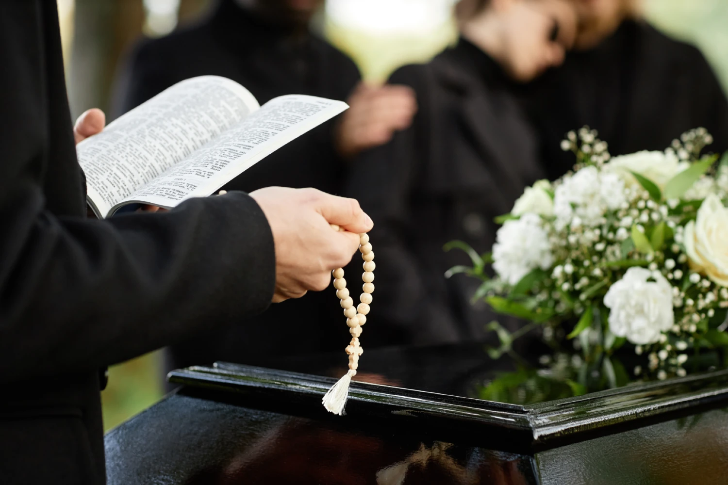 Person holding prayer beads and a book next to a casket with white flowers, attended by mourners in black.