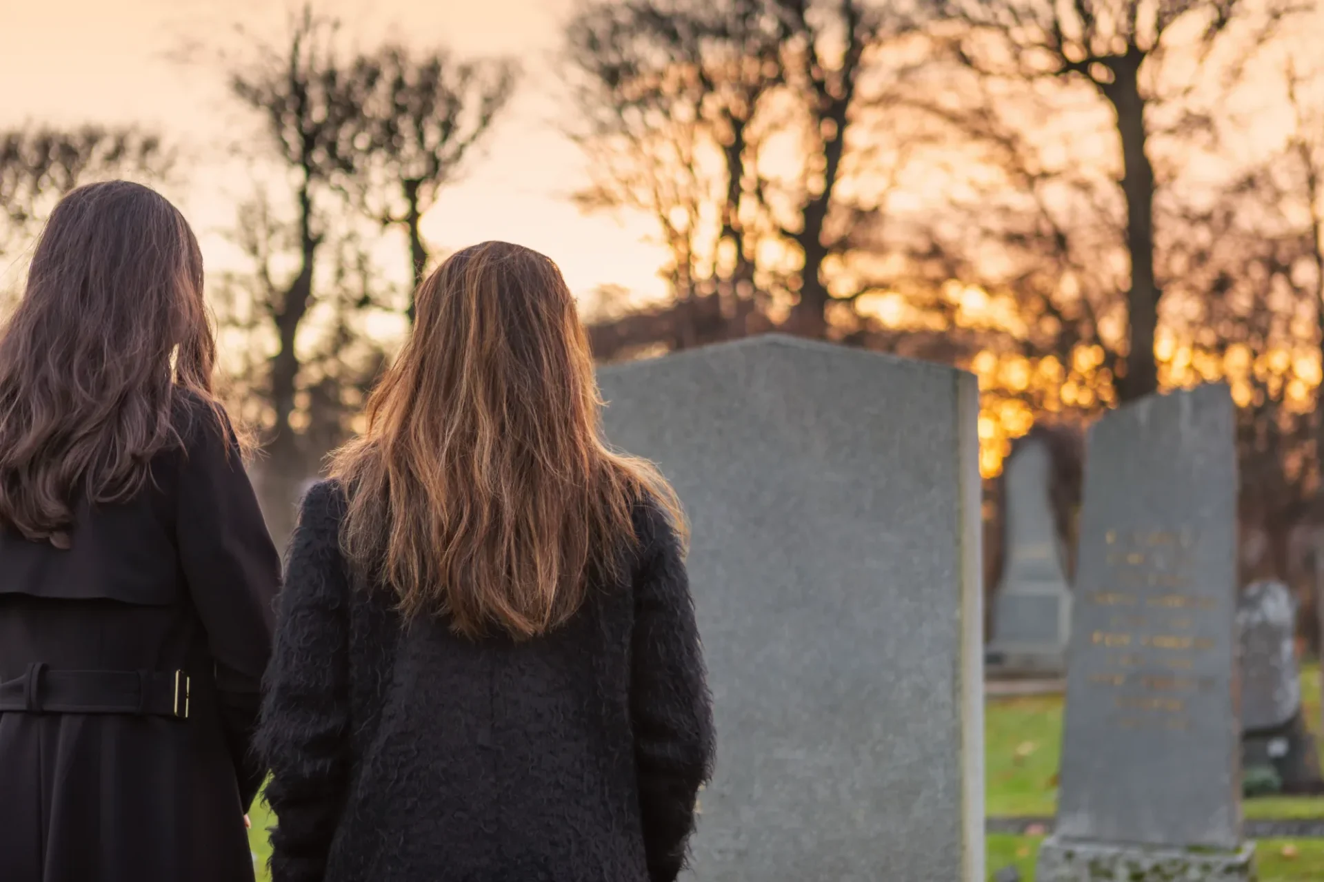 Two people in dark coats stand in front of a gray tombstone at a cemetery, with a sunset and trees in the background.