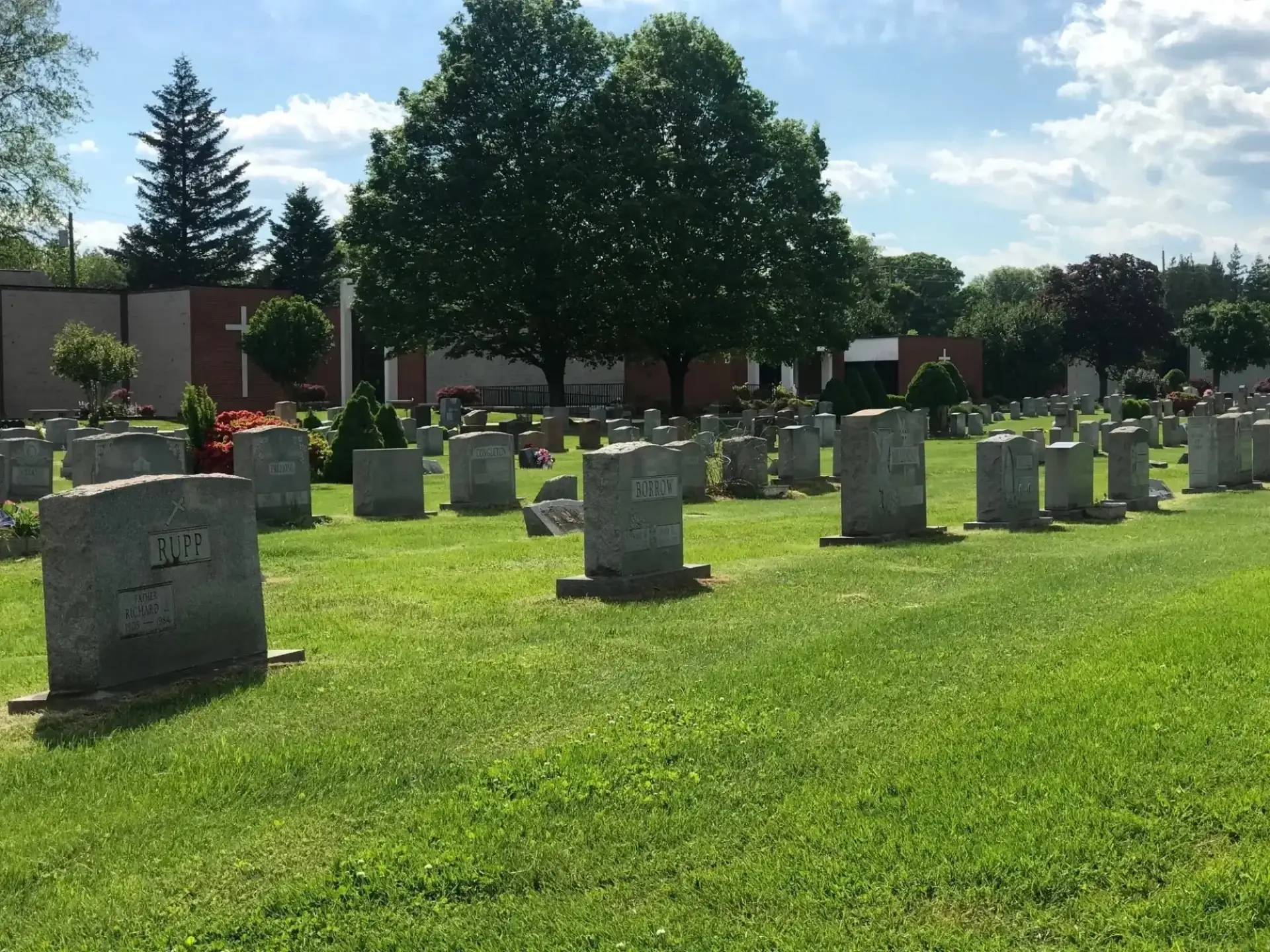 A green cemetery with stone headstones arranged in rows, surrounded by trees under a partly cloudy sky.