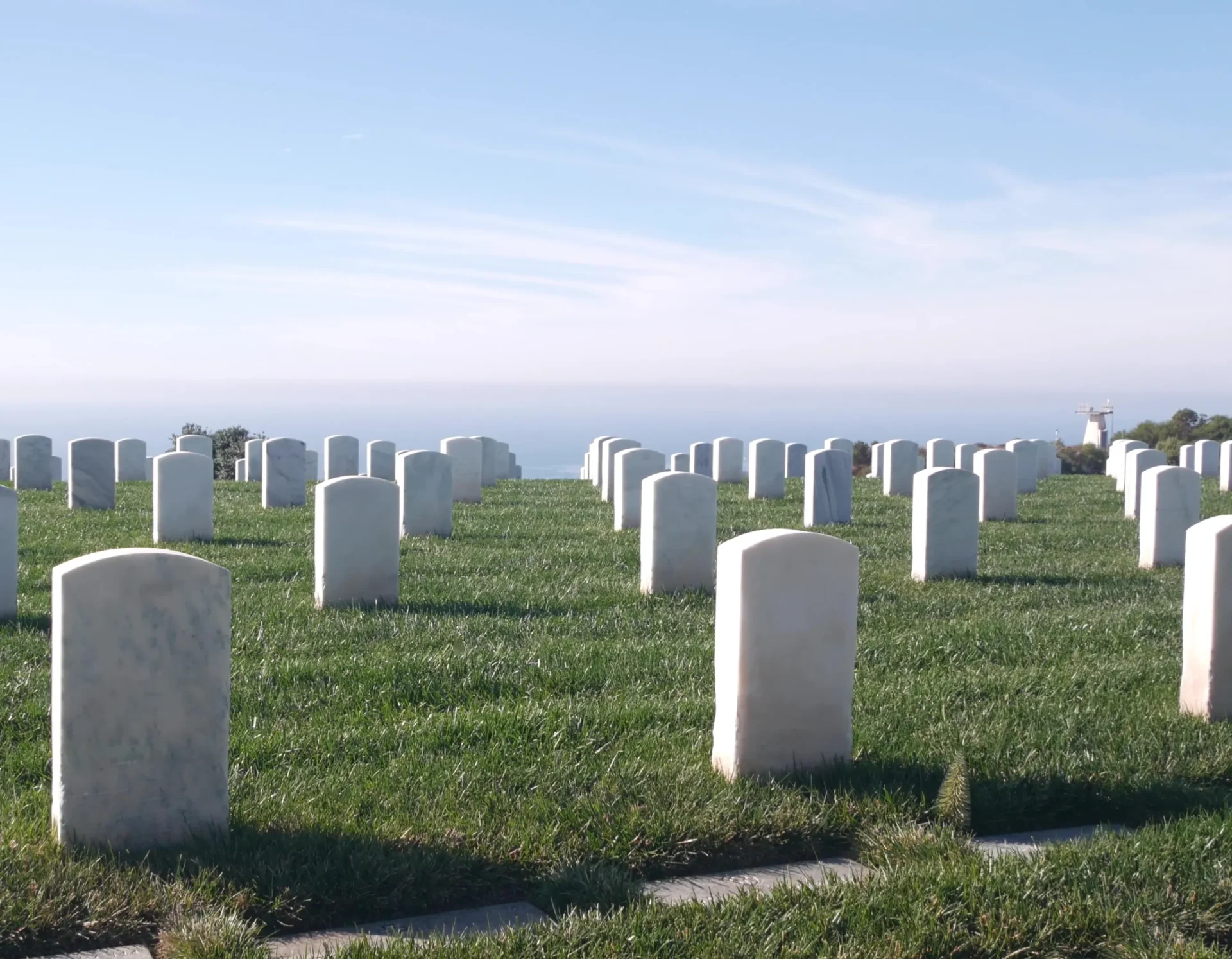 Rows of white tombstones in a cemetery on a clear day, with green grass and a distant view of the ocean.