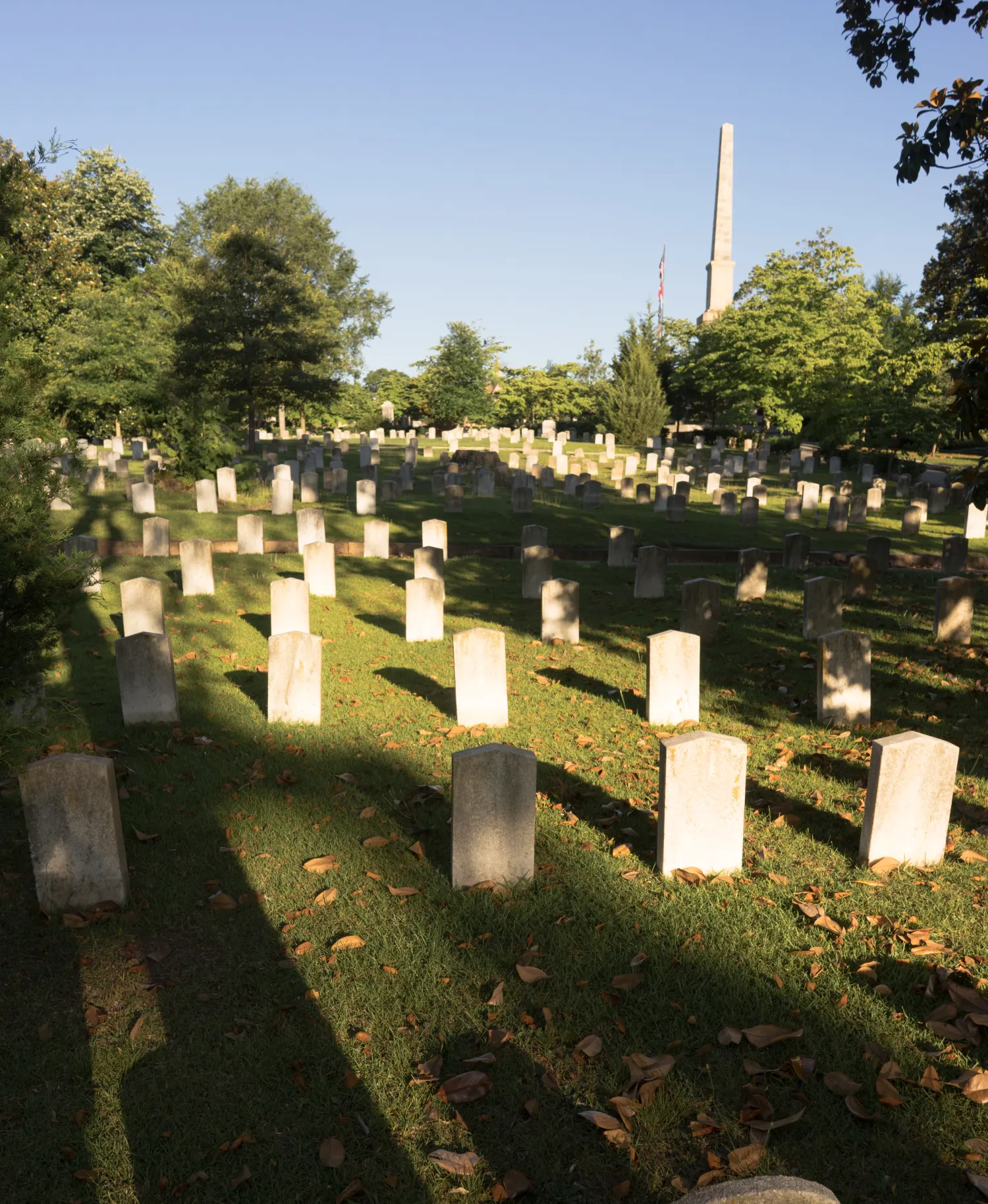 A cemetery with numerous grave markers is shown under a clear blue sky, with a tall obelisk and trees in the background. Shadows from taller objects fall across the grass.