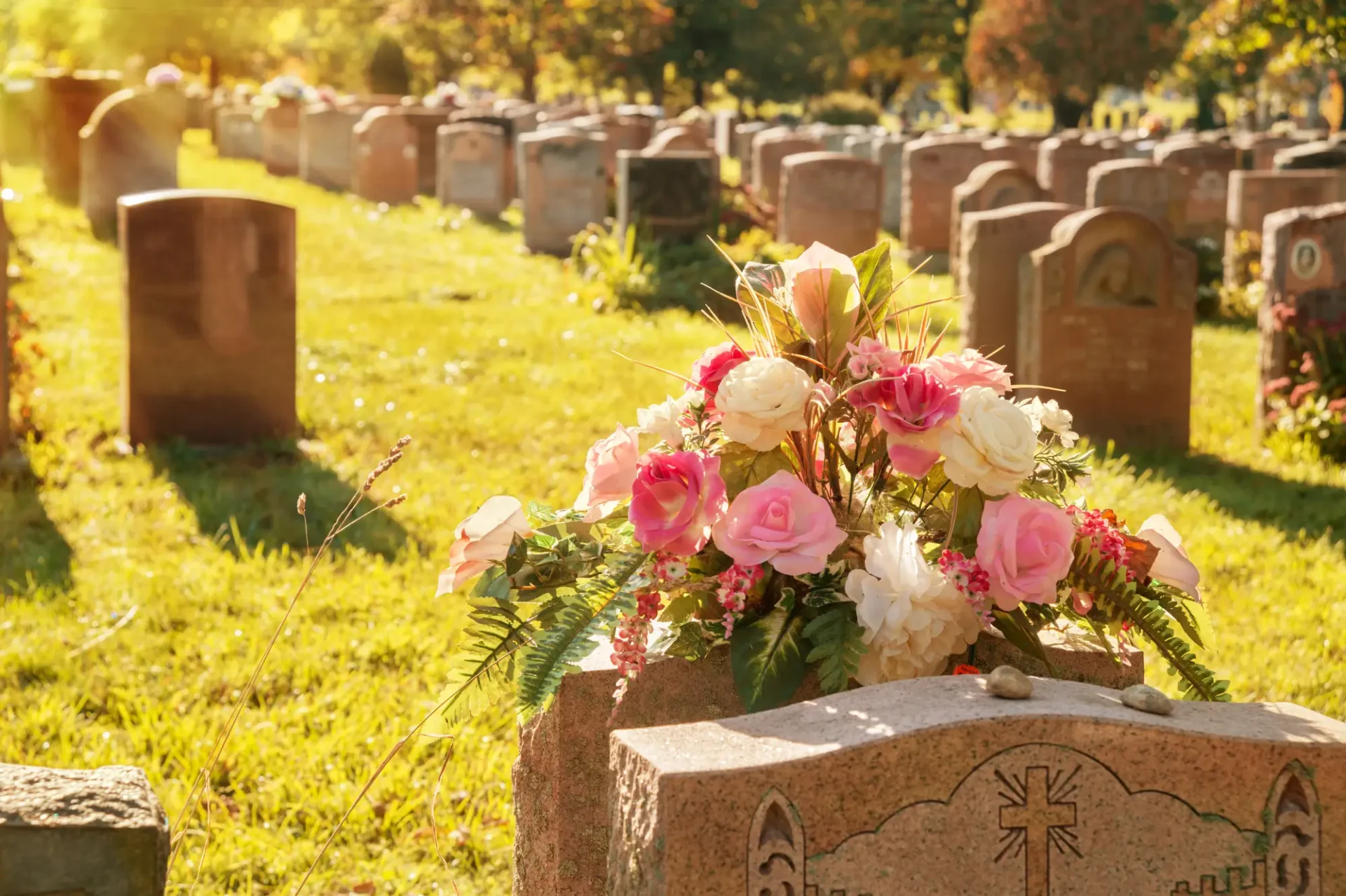 A cemetery with rows of headstones in sunlight. A bouquet of pink and white flowers is placed on a gravestone in the foreground.